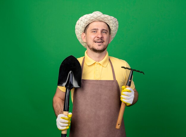 Young gardener wearing jumpsuit and hat in working gloves holding mini rake and shovel looking at front smiling with happy face standing over green wall