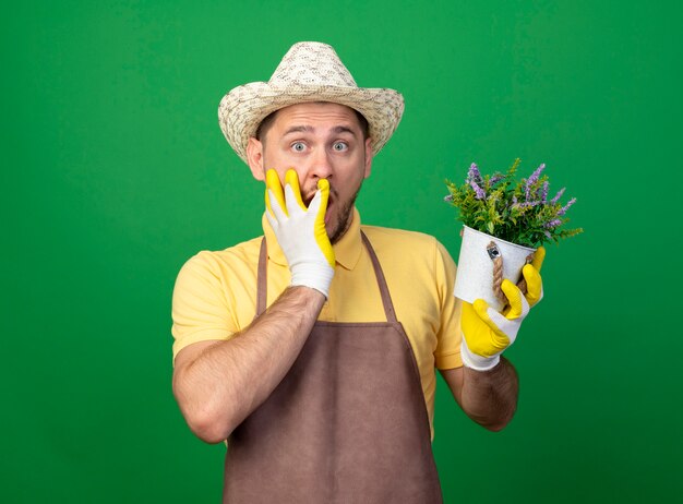 Young gardener wearing jumpsuit and hat in working gloves holding potted plant looking at front beingshocked covering mouth with hand standing over green wall