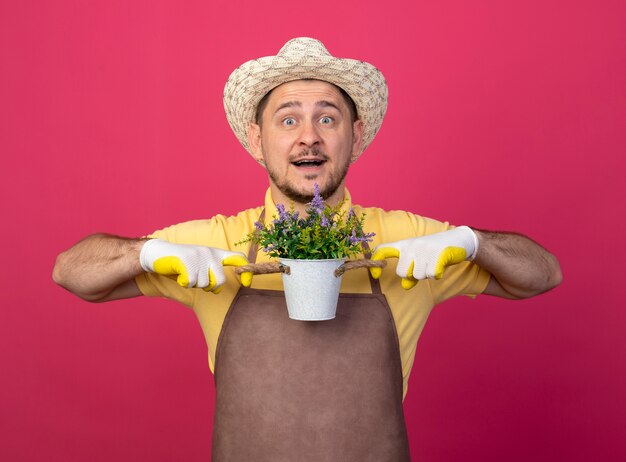 Young gardener wearing jumpsuit and hat in working gloves holding potted plant looking at front with happy face smiling standing over pink wall