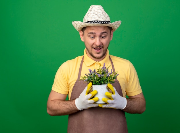 緑の壁の上に立って驚いてそれを見て鉢植えの植物を保持している作業手袋でジャンプスーツと帽子を身に着けている若い庭師