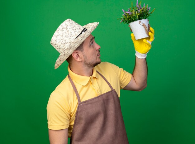 緑の壁の上に立って興味をそそられてそれを見て鉢植えの植物を保持している作業手袋でジャンプスーツと帽子を身に着けている若い庭師