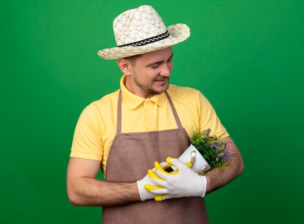 緑の壁の上に立って笑顔でそれを見ている鉢植えの植物を保持している作業手袋でジャンプスーツと帽子を身に着けている若い庭師