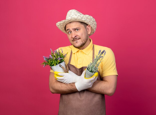 Young gardener wearing jumpsuit and hat in working gloves holding potted plants looking at front confused standing over pink wall