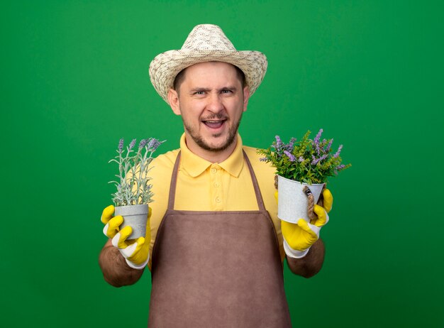 Young gardener wearing jumpsuit and hat in working gloves holding potted plants looking at front smiling cheerfully standing over green wall