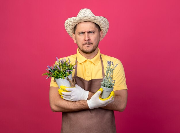 Young gardener wearing jumpsuit and hat in working gloves holding potted plants looking at front with serious face standing over pink wall