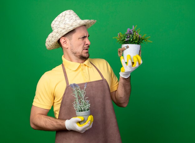 Young gardener wearing jumpsuit and hat in working gloves holding potted plants looking at them being confused and displeased standing over green wall