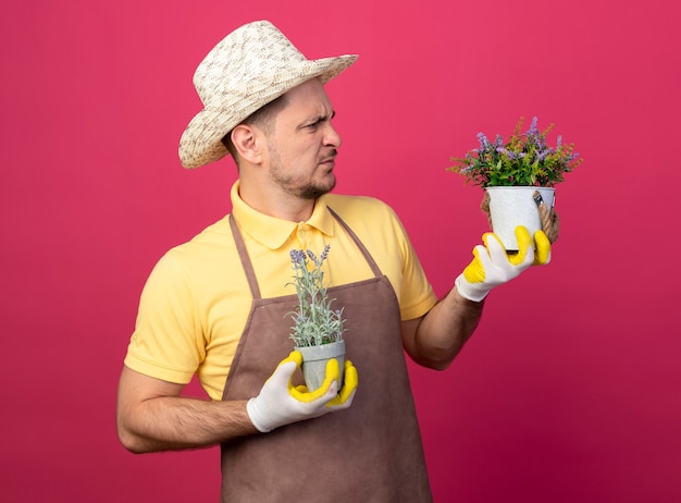 Young gardener wearing jumpsuit and hat in working gloves holding potted plants looking at them being confused and displeased standing over pink wall