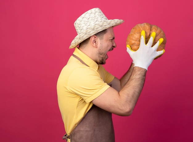 ピンクの壁に攻撃的な表情で叫んで横に立っているカボチャを保持している作業手袋でジャンプスーツと帽子を身に着けている若い庭師