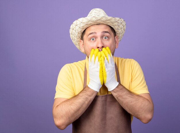 Young gardener wearing jumpsuit and hat in working gloves looking at front being shocked covering mouth with hands standing over purple wall