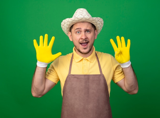 Young gardener wearing jumpsuit and hat in working gloves showing and pointing up with fingers number nine smiling cheerfully standing over green wall