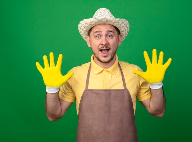 Young gardener wearing jumpsuit and hat in working gloves showing and pointing up with fingers number ten happy and surprised standing over green wall