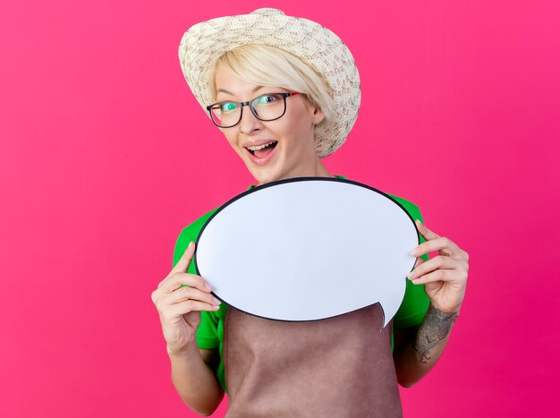 Young gardener woman with short hair in apron and hat holding blank speech bubble sign looking at camera smiling with happy face standing over pink background