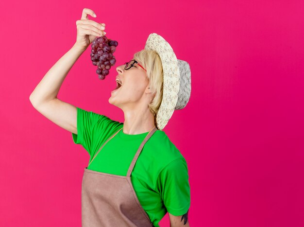 Young gardener woman with short hair in apron and hat holding bunch of grapes