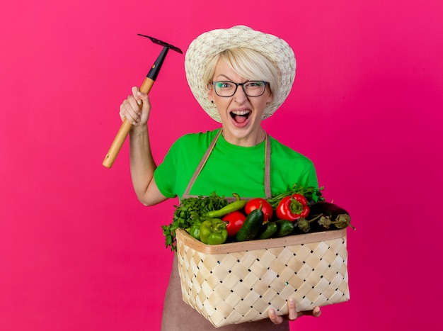 Молодая женщина-садовник с короткими волосами в фартуке и шляпе держит ящик, полный овощей