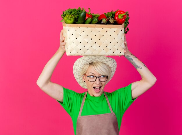 Young gardener woman with short hair in apron and hat holding crate full of vegetables