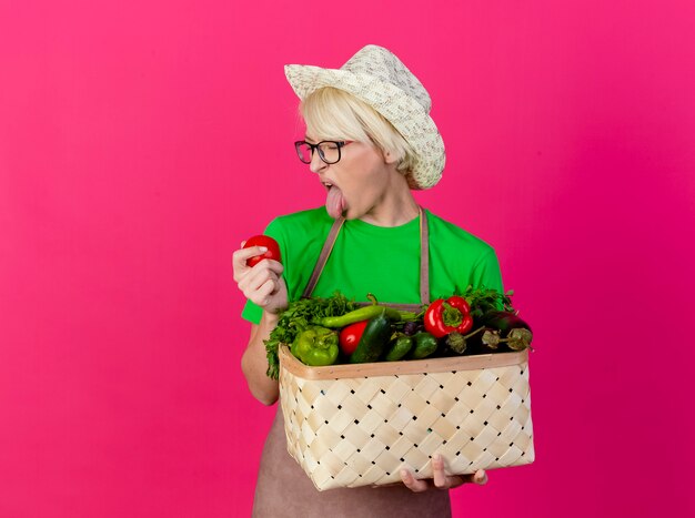 Young gardener woman with short hair in apron and hat holding crate full of vegetables