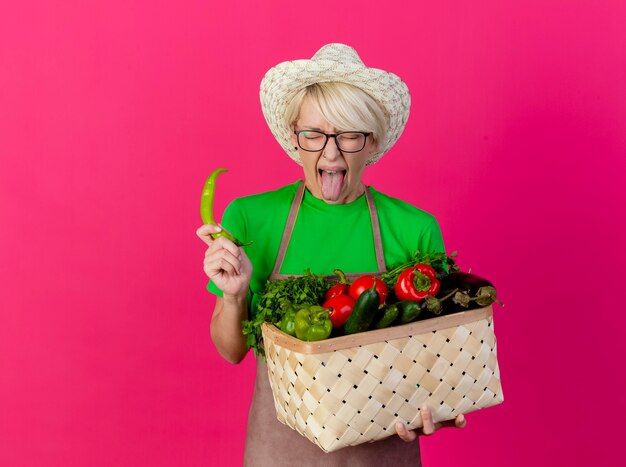 Giovane giardiniere donna con i capelli corti in grembiule e cappello tenendo la cassa piena di verdure