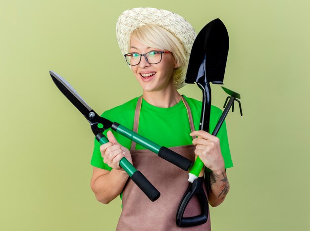 Young gardener woman with short hair in apron and hat holding gardening equipments looking at camera with smile on happy face standing over light background