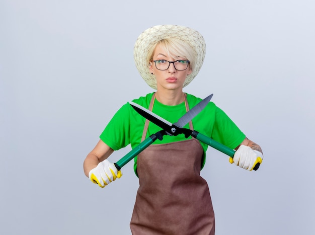 Young gardener woman with short hair in apron and hat holding hedge clippers with serious face