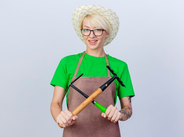 Young gardener woman with short hair in apron and hat holding mattock and mini rake crossing hands smiling cheerfully
