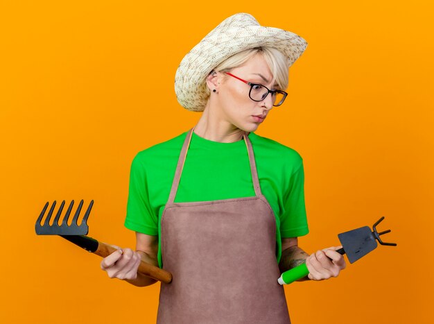 Young gardener woman with short hair in apron and hat holding mattock and mini rake looking confused trying to make choice standing over orange background