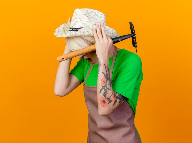 Young gardener woman with short hair in apron and hat holding mini rake holding her head with annoyed expression standing over orange background