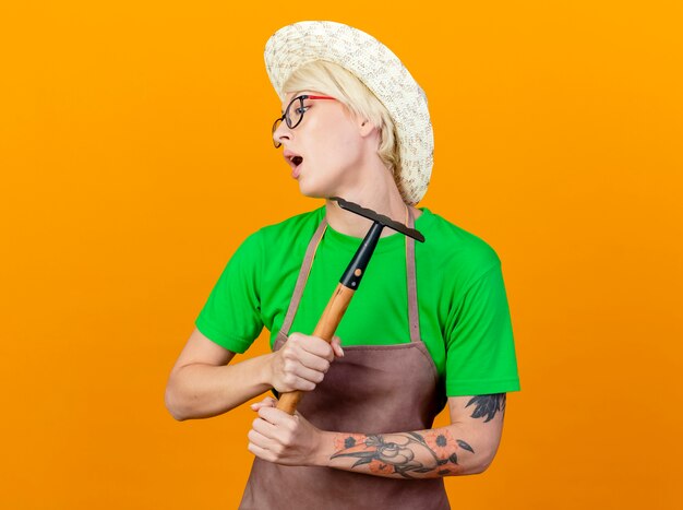 Young gardener woman with short hair in apron and hat holding mini rake looking aside confused standing over orange background