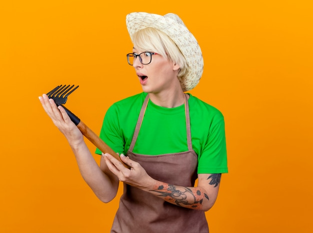 Young gardener woman with short hair in apron and hat holding mini rake looking at it amazed standing over orange background