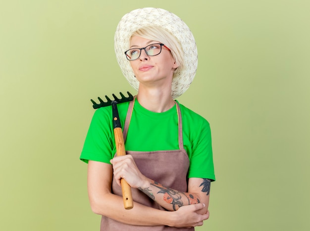 Young gardener woman with short hair in apron and hat holding mini rake looking up with skeptic smile on face standing over light background