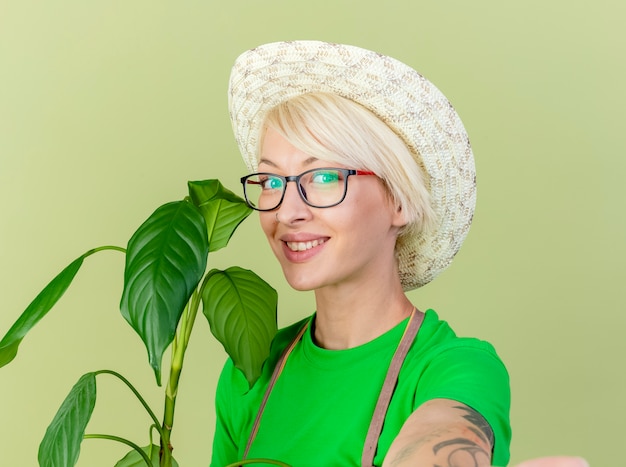 앞치마에 짧은 머리를 가진 젊은 정원사 여자와 밝은 배경 위에 서있는 얼굴에 미소로 카메라를보고 식물을 들고 모자