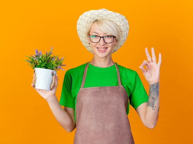 Young gardener woman with short hair in apron and hat holding potted plant looking at camera smiling with happy face showing ok sign standing over orange background