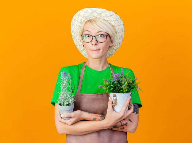 エプロンの短い髪と鉢植えの植物を保持している帽子を持つ若い庭師の女性は、オレンジ色の背景の上に立っている懐疑的な笑顔でカメラを見て