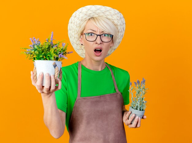 Young gardener woman with short hair in apron and hat holding potted plants looking at camera being amazed and surprised standing over orange background