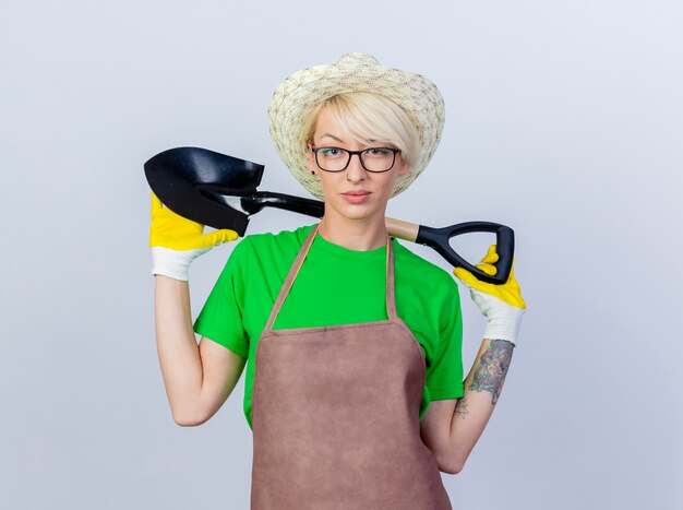 Young gardener woman with short hair in apron and hat holding shovel  with serious confident expression