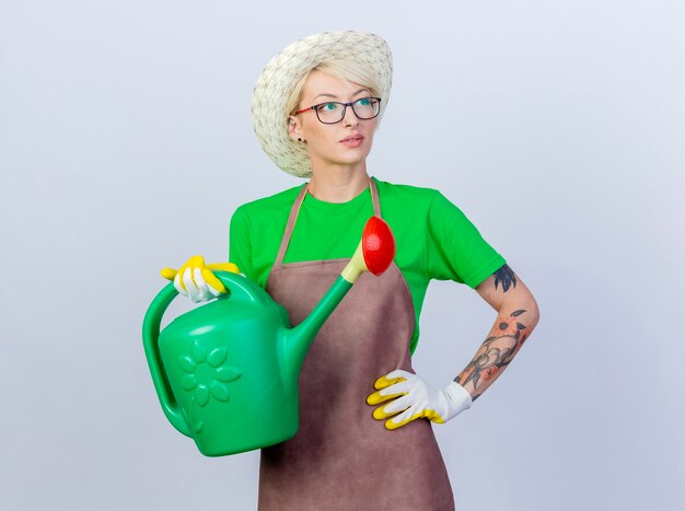 Young gardener woman with short hair in apron and hat holding watering can looking aside puzzled and thinking