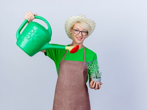 Young gardener woman with short hair in apron and hat holding watering can and potted plant smiling with happy face