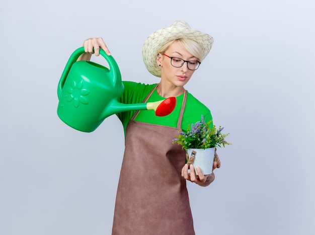 Young gardener woman with short hair in apron and hat holding watering can and potted plant watering it looking confident