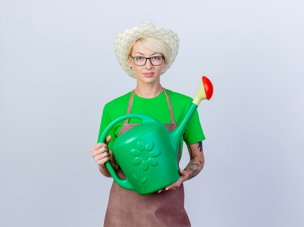 Young gardener woman with short hair in apron and hat holding watering can with smile on face