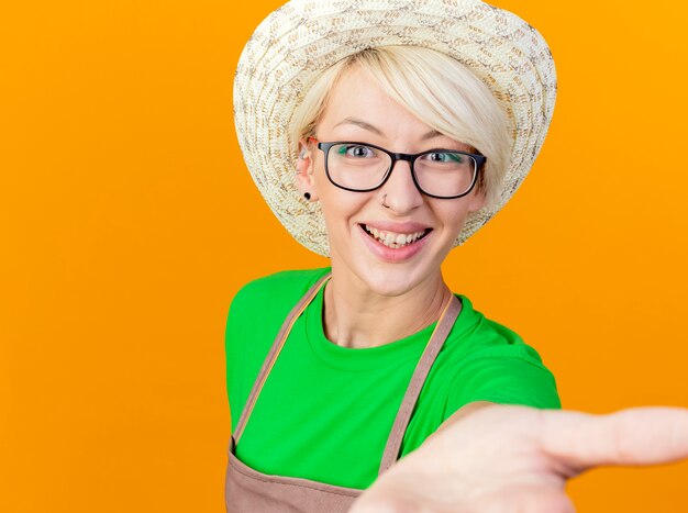 Young gardener woman with short hair in apron and hat looking at camera with arm out smiling cheerfully standing over orange background