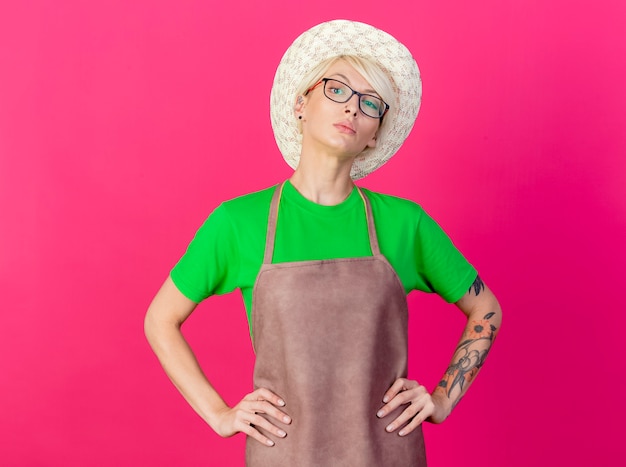 Foto gratuita giovane giardiniere donna con i capelli corti in grembiule e cappello guardando fiducioso con le braccia all'anca