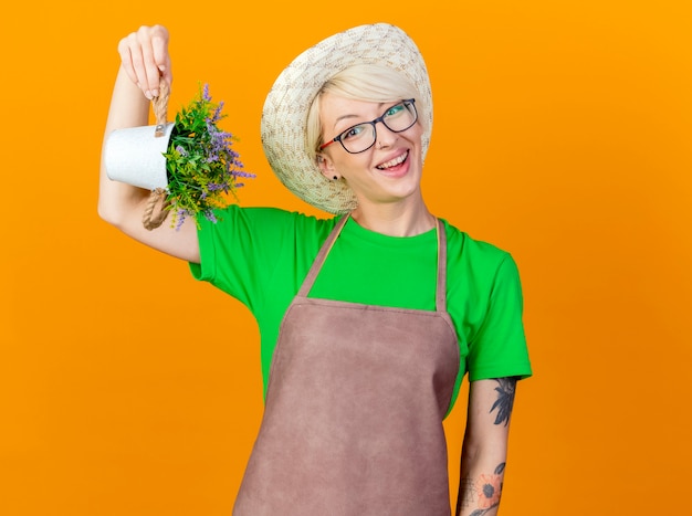 Young gardener woman with short hair in apron and hat showing potted plant looking at camera smiling with happy face standing over orange background