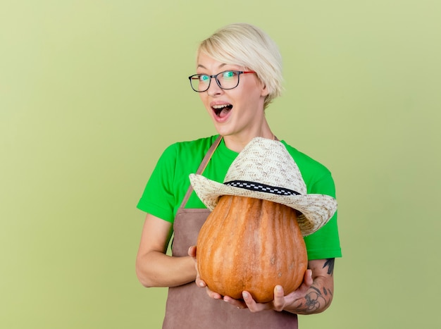 Young gardener woman with short hair in apron holding pumpkin with hat on it looking at camera with smile on face standing over light background