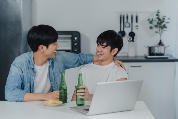 Young Gay couple drink beer while using computer laptop at modern home. Asian LGBTQ men happy relax fun using technology play social media together while sitting table in kitchen at house .