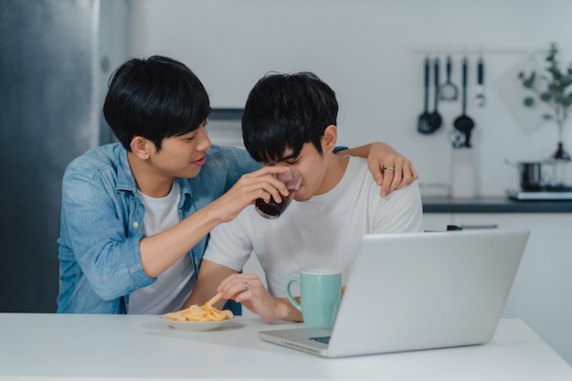 Young Gay couple feeding food and snack using computer laptop at modern home. Asian LGBTQ men happy relax fun using technology social media together while sitting table in kitchen at house .