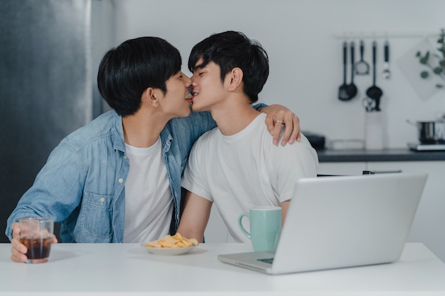 若い​同性愛者​の​カップル​が​現代​の​家​で​コンピューター​の​ラップ​トップ​を​使用し​ながら​キスします​。​アジア​の​lgbtq​男性​は​、​家​の​台所​で​テーブル​に​座って​ソーシャルメディア​を​一緒​に​再生する​技術​を​使用して​楽しい​リラックス​を​楽しんでいます​。