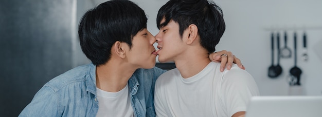 Free photo young gay couple kissing while using computer laptop at modern home. asian lgbtq men happy relax fun using technology play social media together while sitting table in kitchen at house .