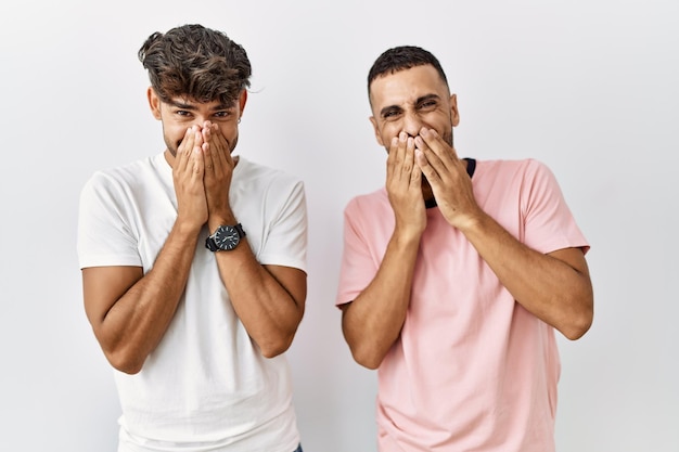 Young gay couple standing over isolated background laughing and embarrassed giggle covering mouth with hands, gossip and scandal concept