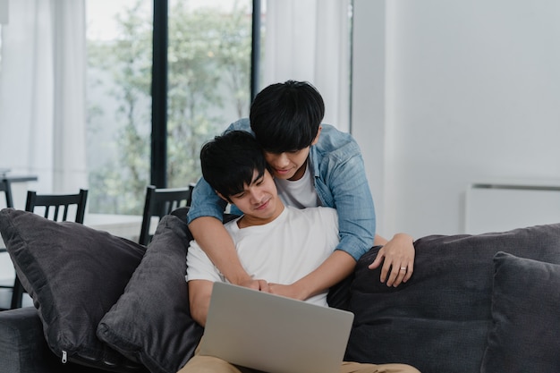 현대 집에서 컴퓨터 노트북을 사용하는 젊은 게이 커플. 아시아 LGBTQ + 남성은 집에서 거실에 소파에 누워있는 동안 인터넷에서 함께 영화를보고 기술을 사용하여 즐거운 휴식을 취하십시오.