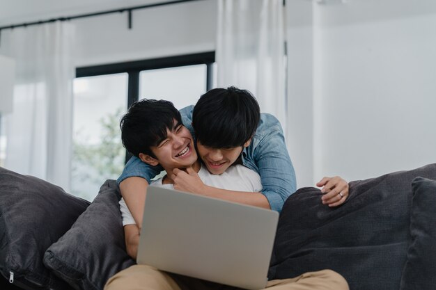 Молодые пары гомосексуалиста используя компьтер-книжку компьютера на современном доме. Азиатские LGBTQ + мужчины счастливы расслабиться весело, используя технологии, смотря фильмы в интернете вместе, лежа на диване в гостиной дома.