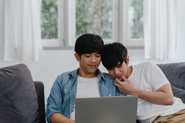 Young Gay couple using computer laptop at modern home. Asian LGBTQ+ men happy relax fun using technology watching movie in internet together while lying sofa in living room at house .